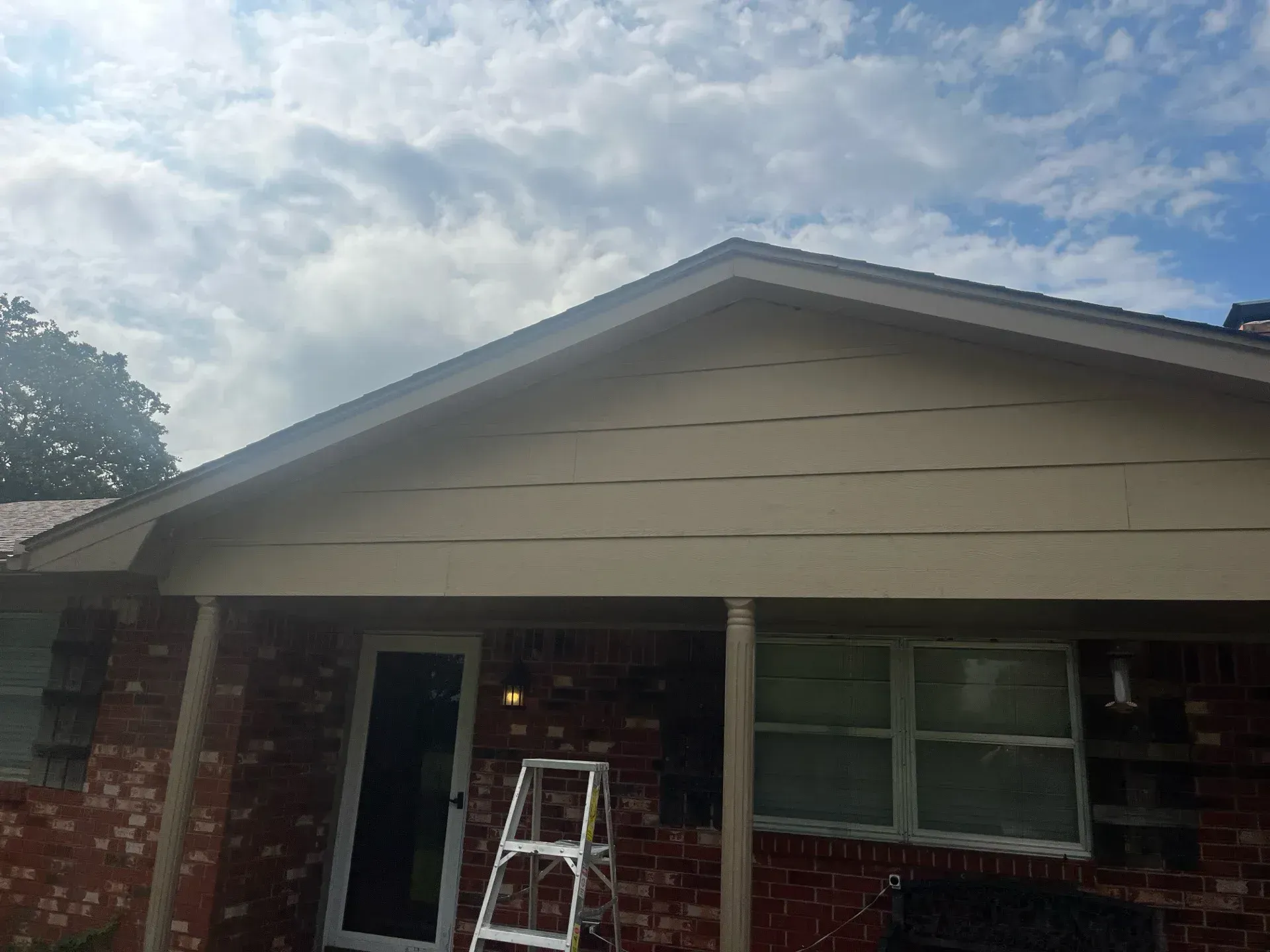 House with red brick exterior, beige siding, and a ladder. Cloudy sky.