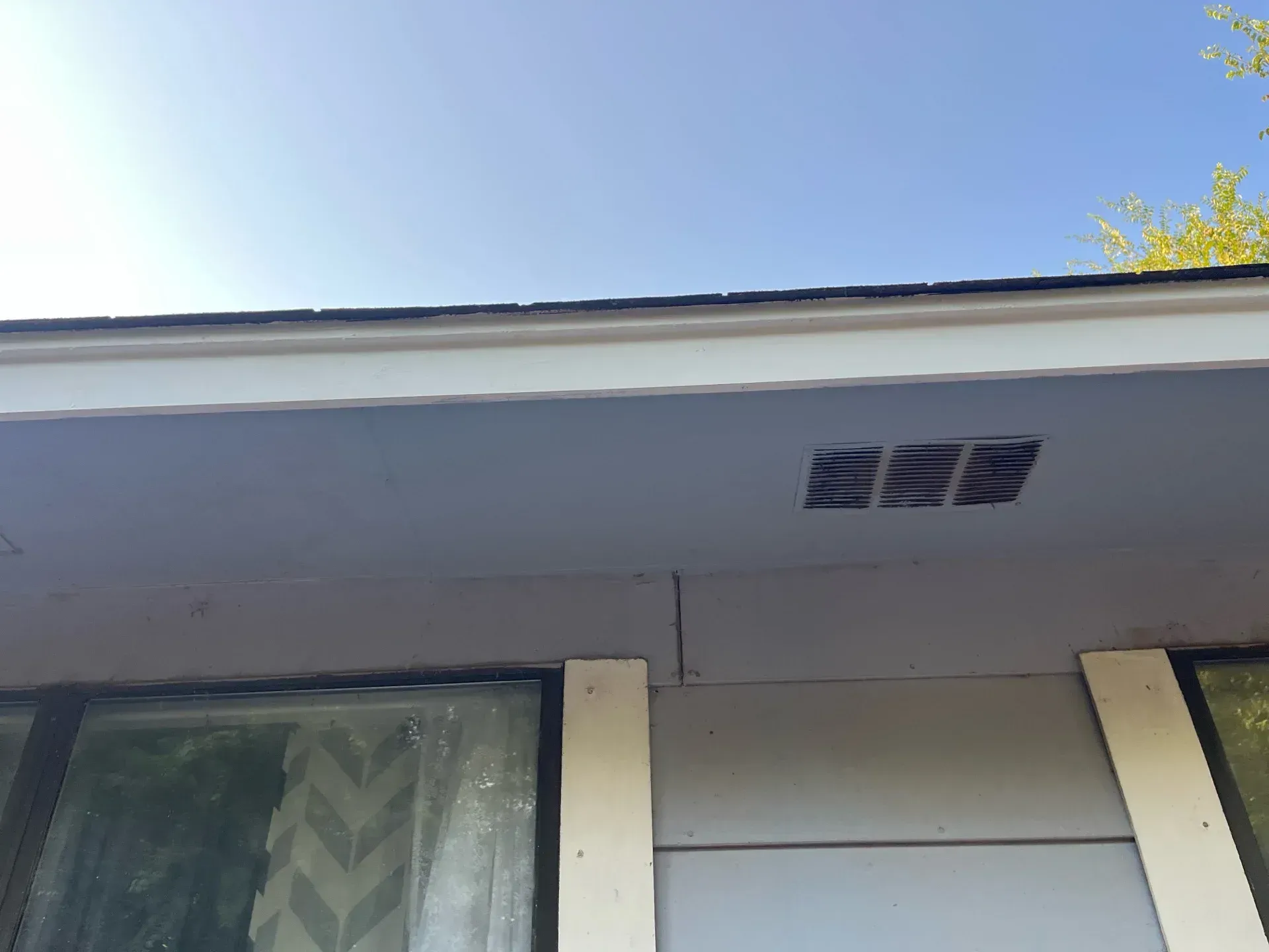 Exterior house wall with soffit vents and a window under a clear blue sky.