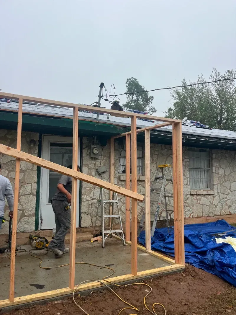 Construction workers building a wooden frame on a home's patio. Gray sky and blue tarps present.