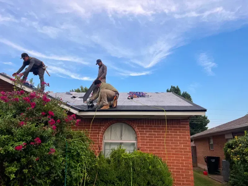 Three workers installing roofing shingles on a residential house under a blue sky.