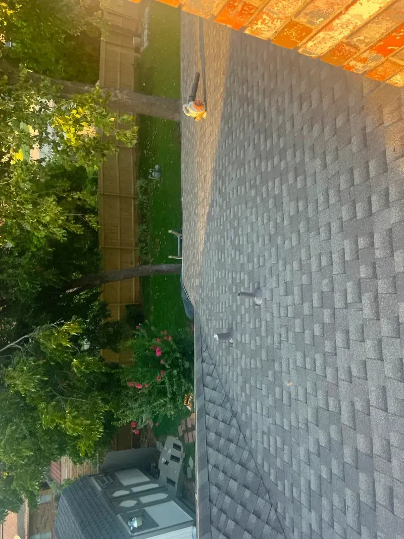 View of a gray shingled roof with a chimney next to a green yard with a wooden fence.