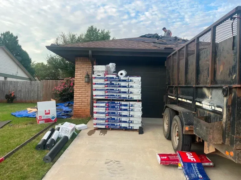 Roofing project: shingles stacked on driveway near a house. A worker is on the roof removing old shingles. A trailer is parked.
