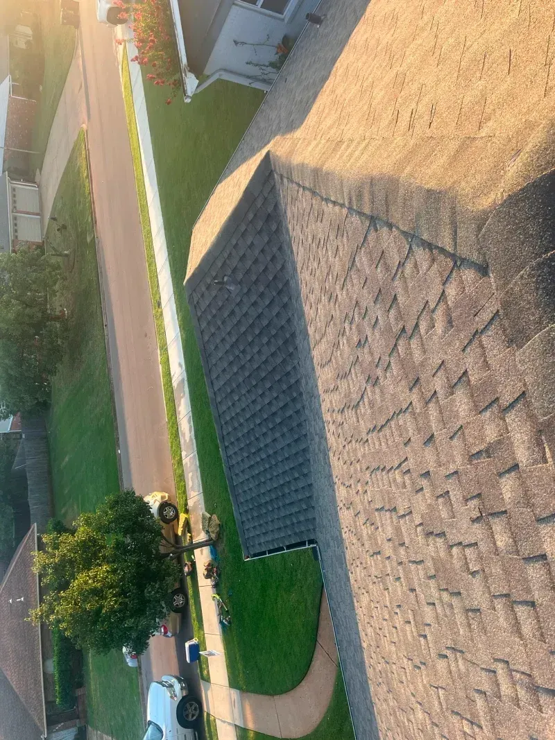Overhead view of a house roof, driveway, street, and greenery, bathed in sunlight.