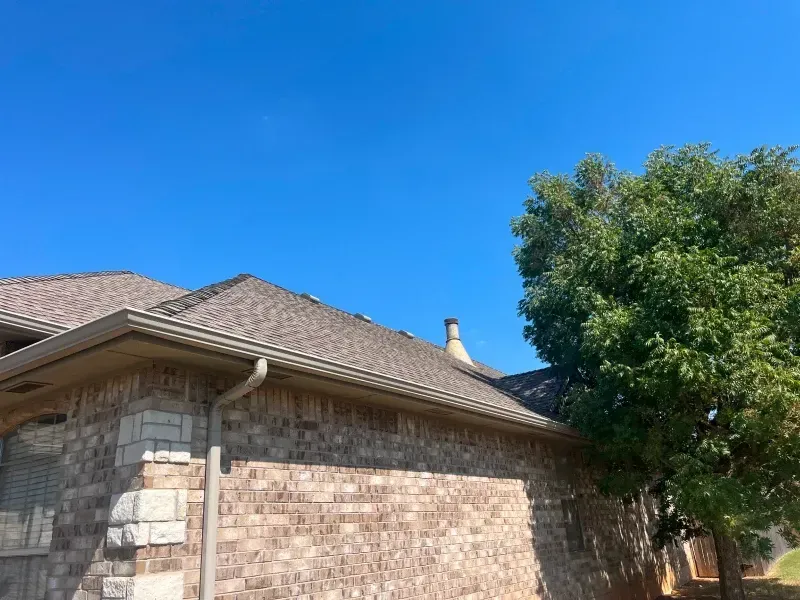 Brick house exterior with brown roof, gutters, and a tree against a blue sky.