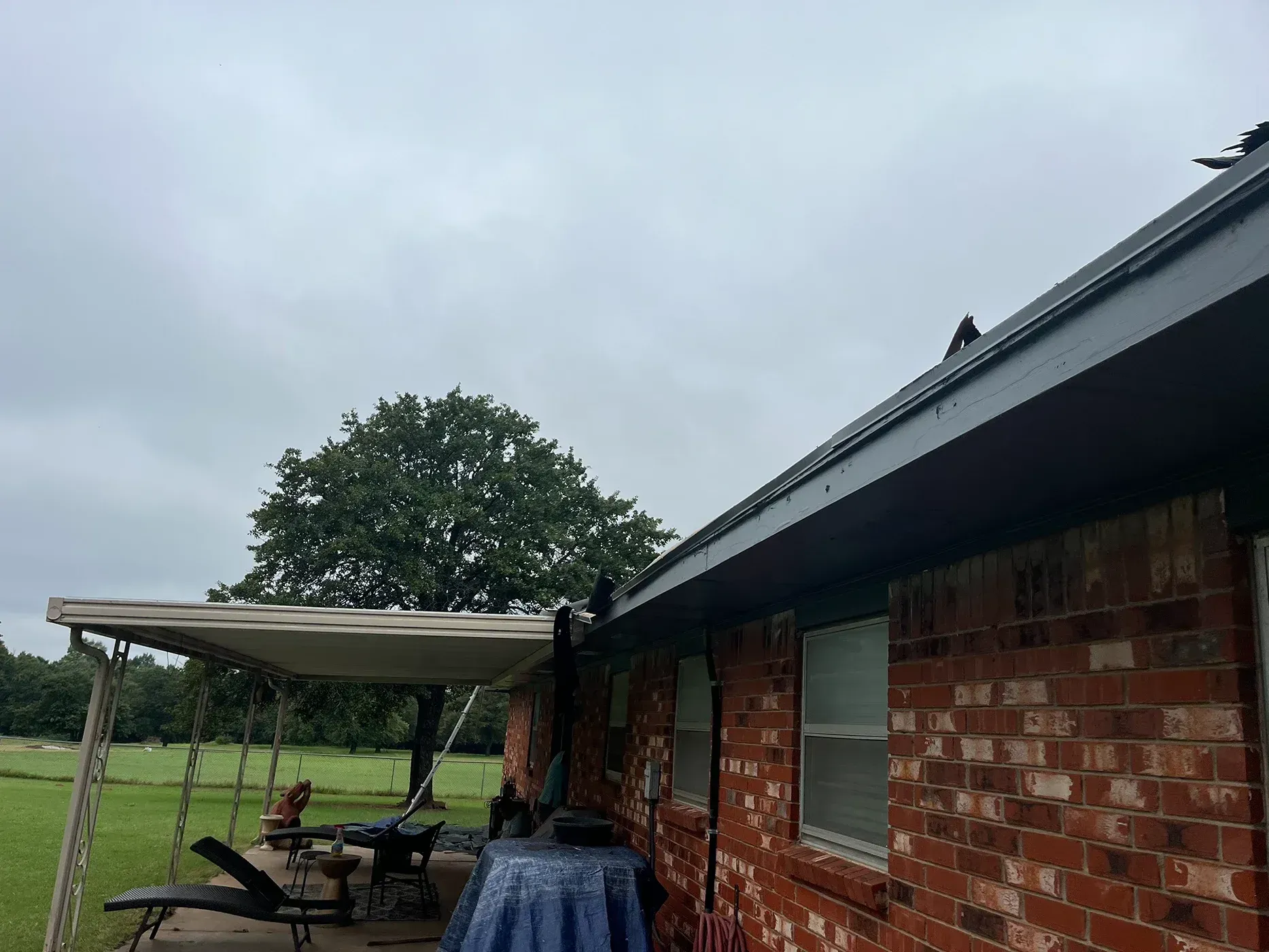 Brick house exterior with a covered patio, overcast sky, and a large tree in the background.