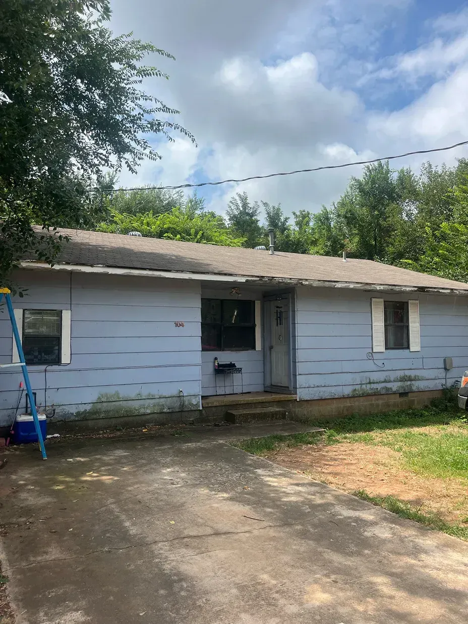 Blue, weathered single-story house with a damaged roof, overgrown foliage, and a cracked driveway.