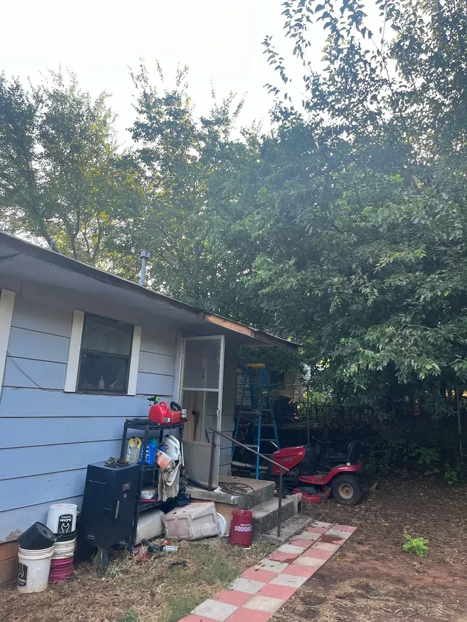 Exterior of a light blue house with a porch and overgrown trees. A red riding mower sits on the side.