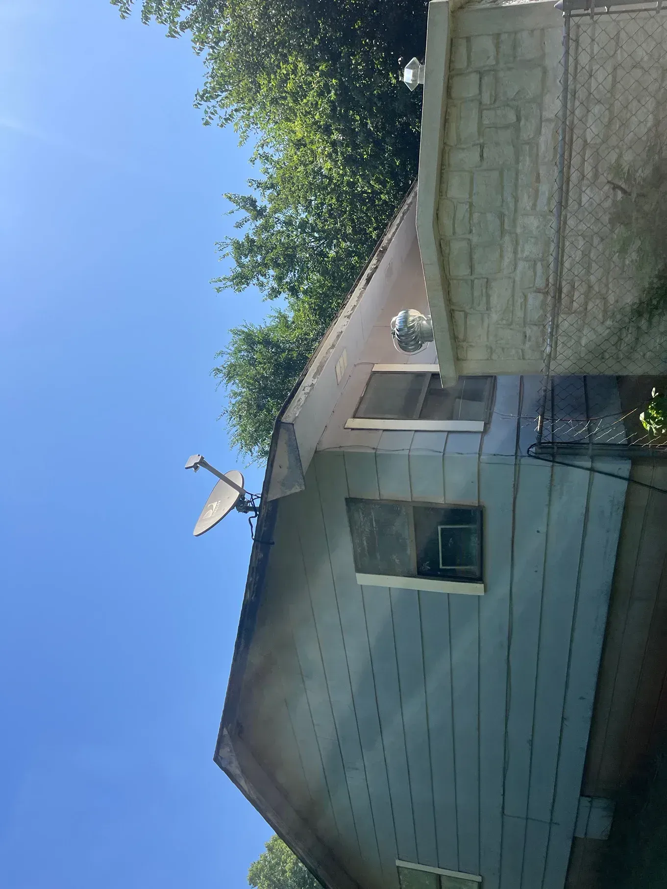 Side of a building with a satellite dish, two windows, and dark discoloration on the roof and siding under a blue sky.