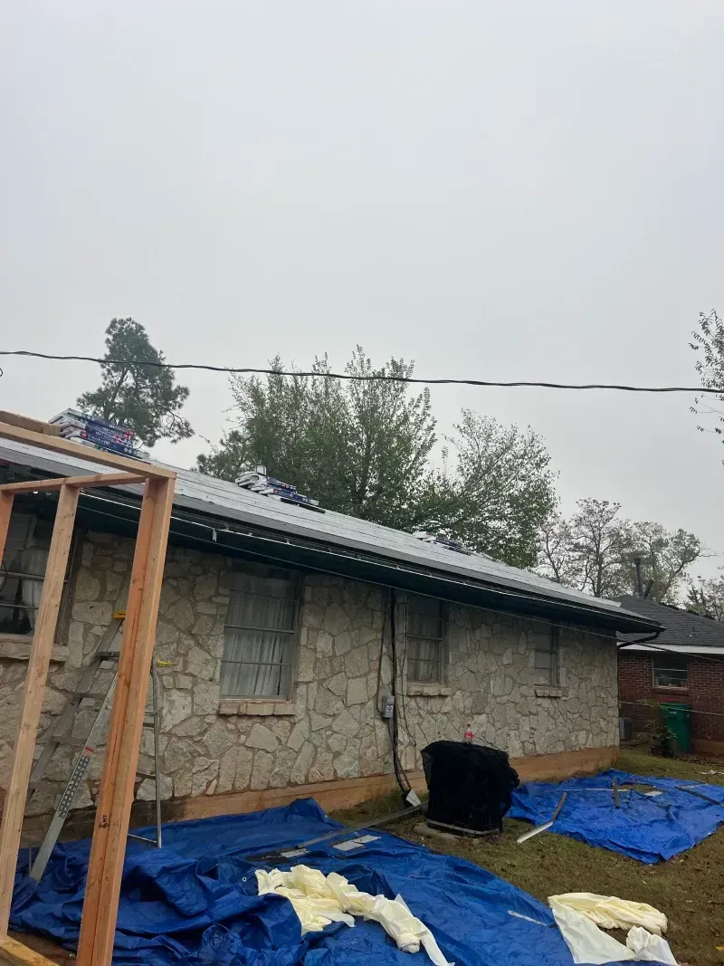 House with stone facade; roof under construction. Blue tarps and wooden frame visible. Overcast day.