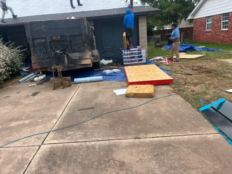 Workers on a roof with a dumpster and materials in a residential driveway.