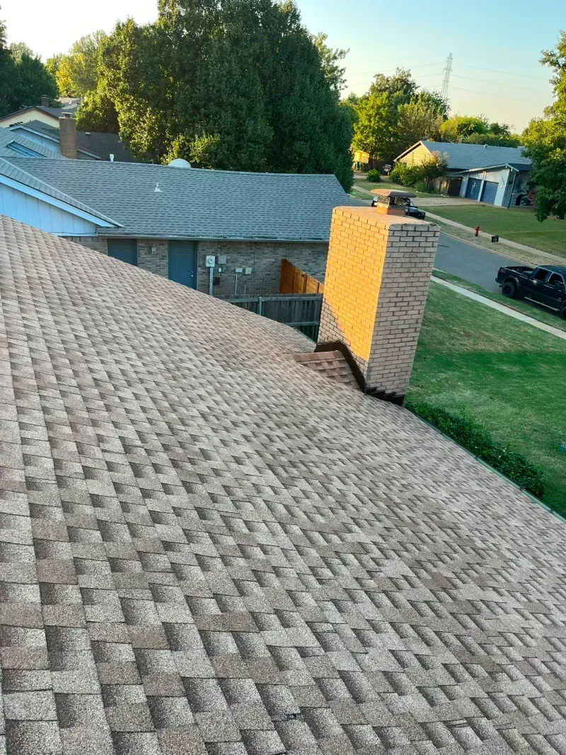 View of a brown shingle roof with a brick chimney and a neighborhood in the background.