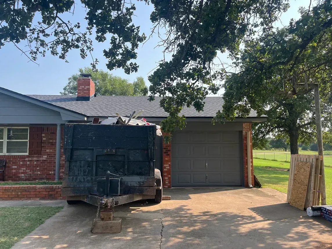 House with a new roof and dumpster in the driveway; a partially open garage door; trees.