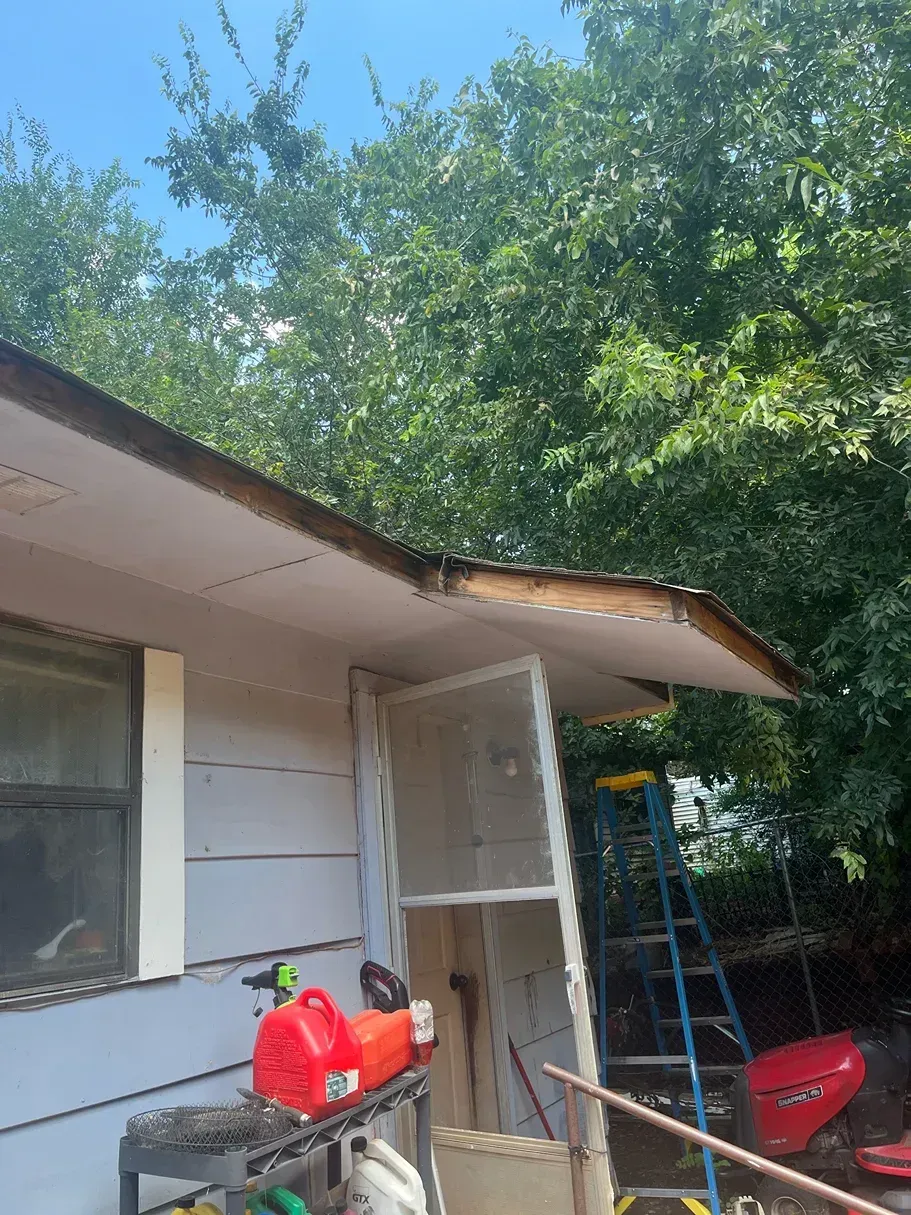 Damaged house roof with exposed wood. Red gas can and tools are on a shelf below. Blue ladder nearby.