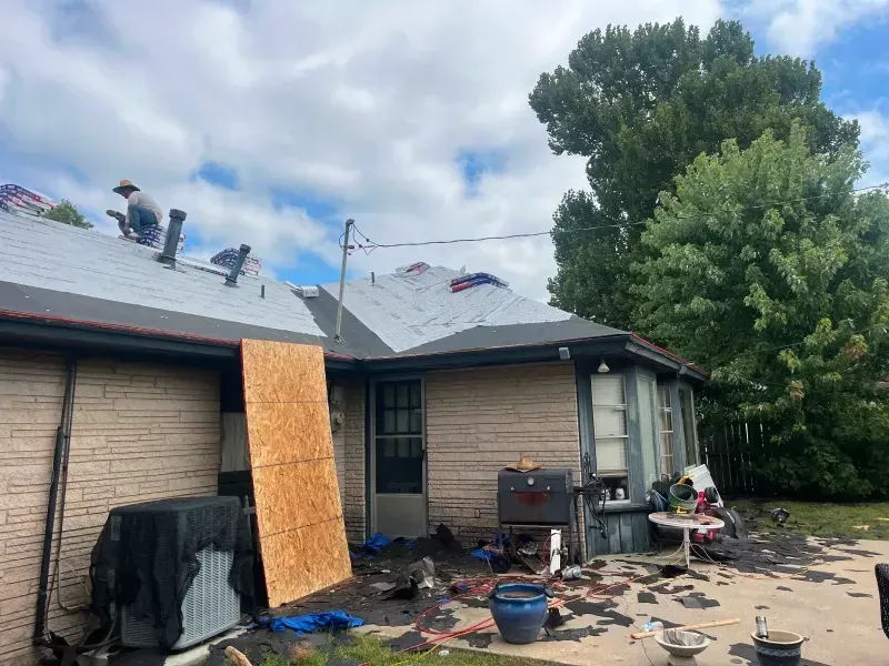 Workers on a roof replacing shingles. House exterior with debris on the ground and a plywood sheet leaning against the wall.