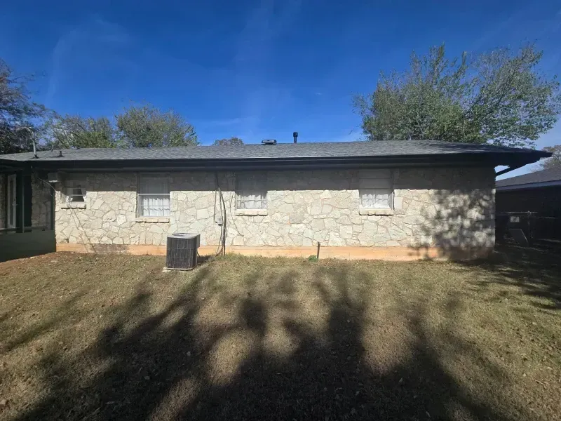 Stone-faced, single-story building with three windows, air conditioning unit, and a grassy yard under a blue sky.