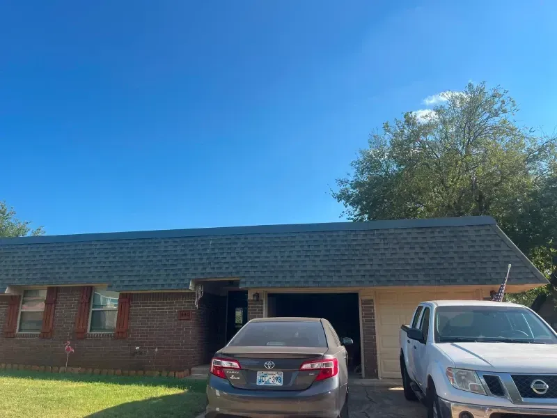 House with green roof, brick and tan exterior, with parked cars and blue sky.