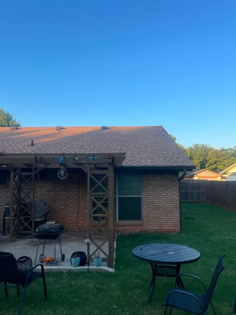 Backyard scene: brick home, patio with grill and table, green lawn, clear blue sky.