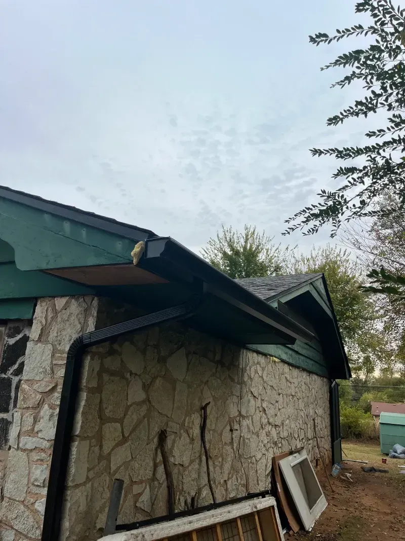 Stone building with green trim and black gutter. Cloudy sky. Board missing from under the gutter.
