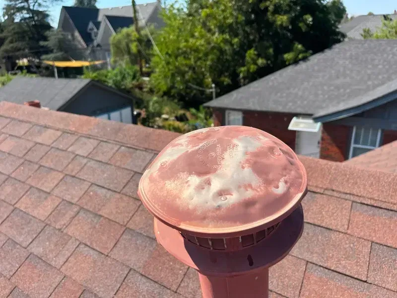 Rusty, domed vent cap on a brown shingled roof, set against a backdrop of houses and trees.