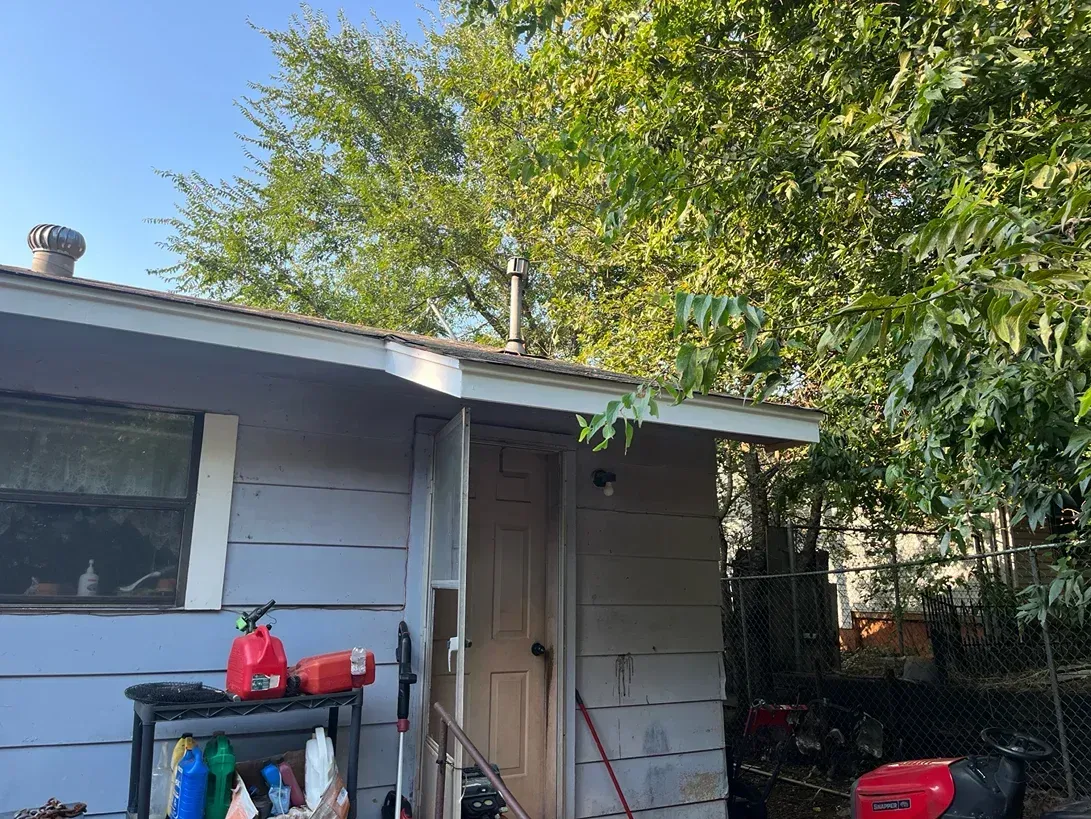 A small gray shed with a chimney, door, and a partially visible window, surrounded by a leafy tree.