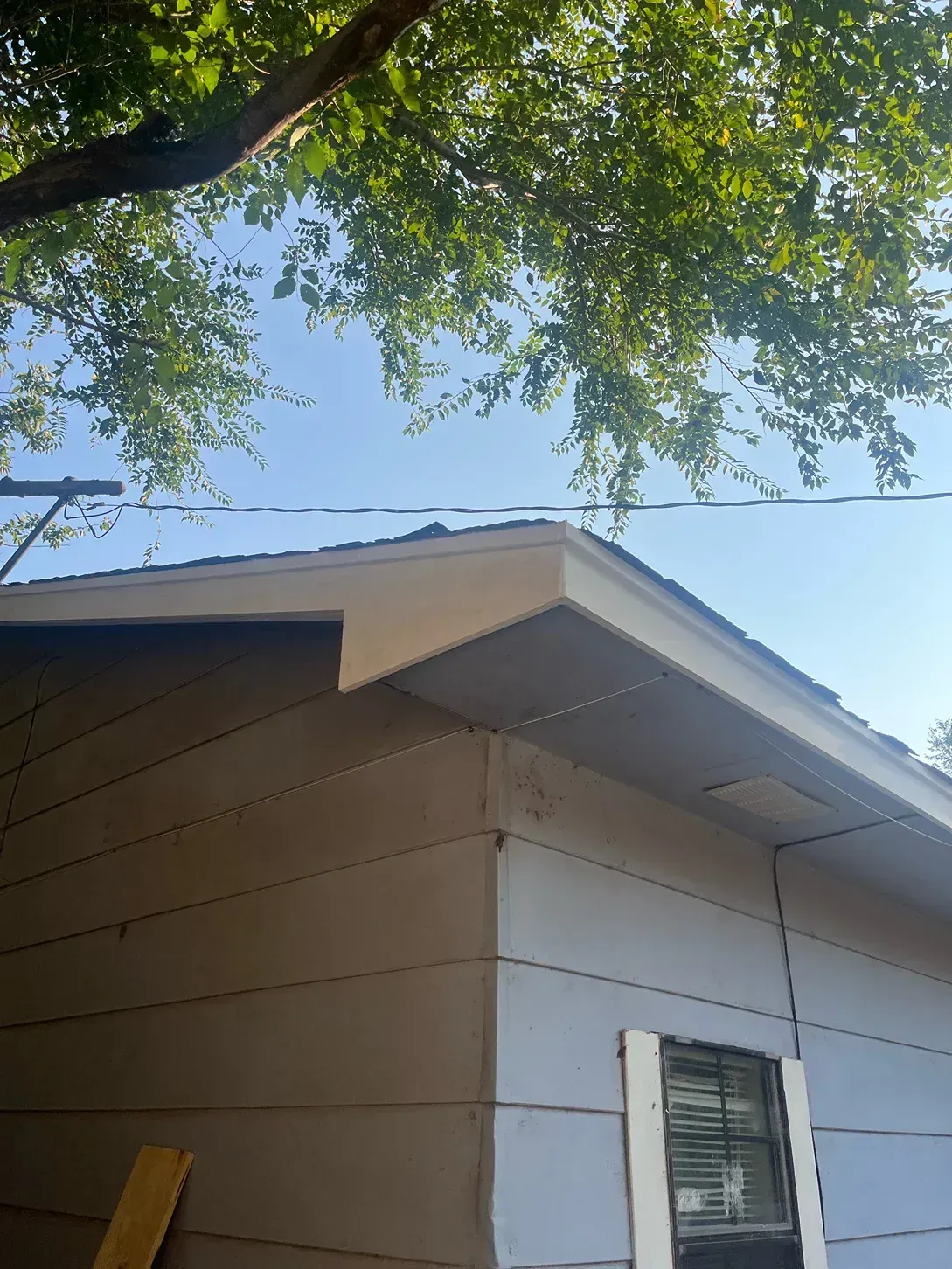 Corner of a light tan house with a beige roof overhang under a bright blue sky. Tree branches are above.
