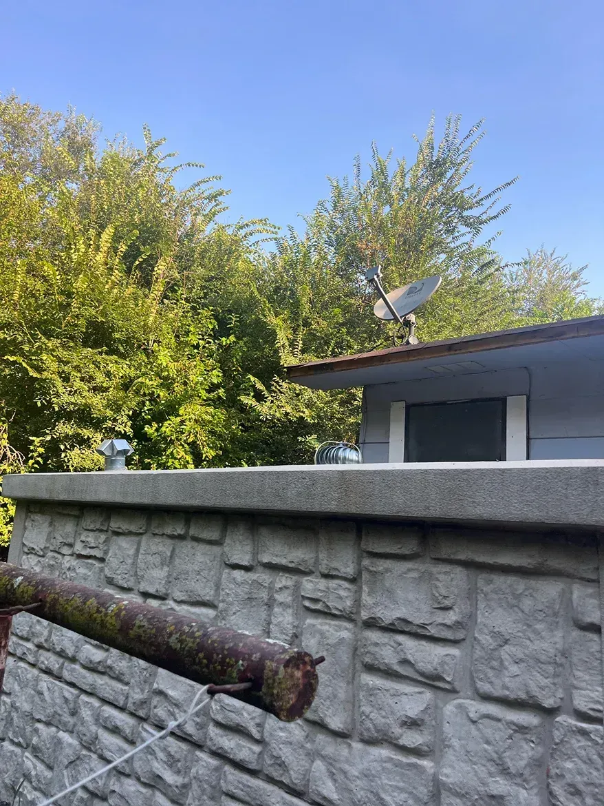 Gray stone wall with a low building and satellite dish against a blue sky with green trees.