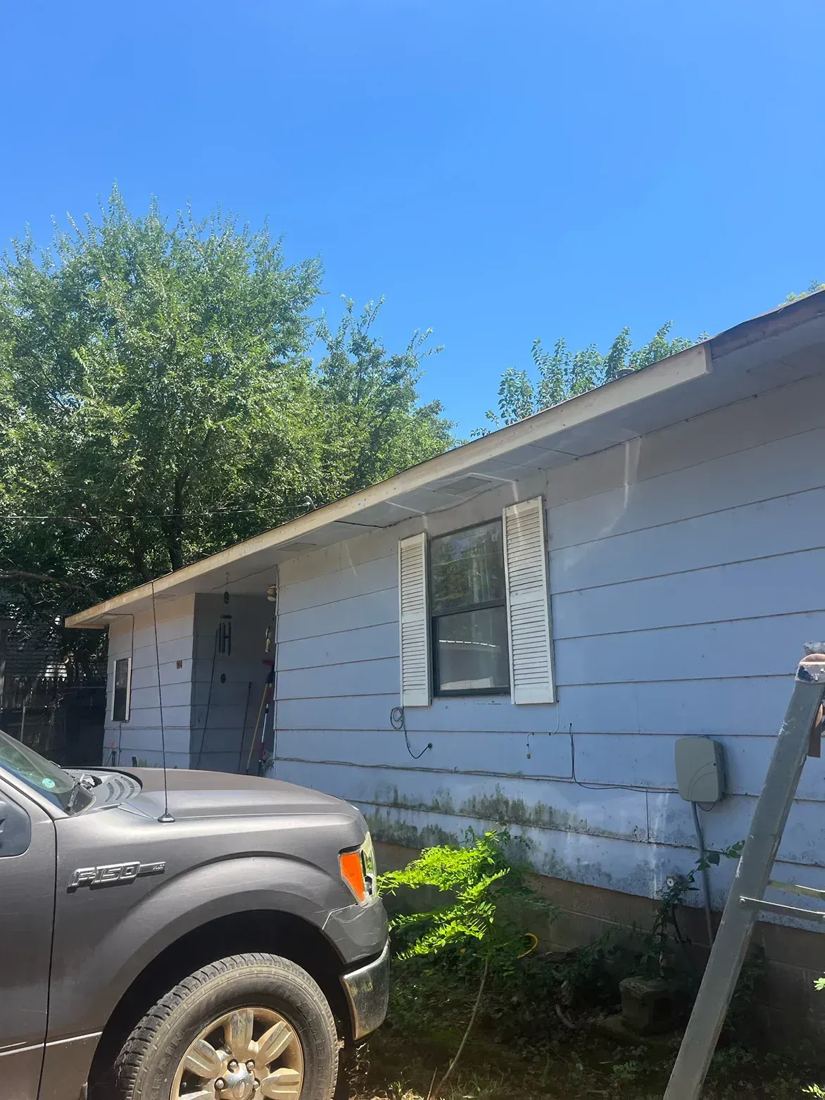 Gray house with white shutters, a truck, and trees under a blue sky.