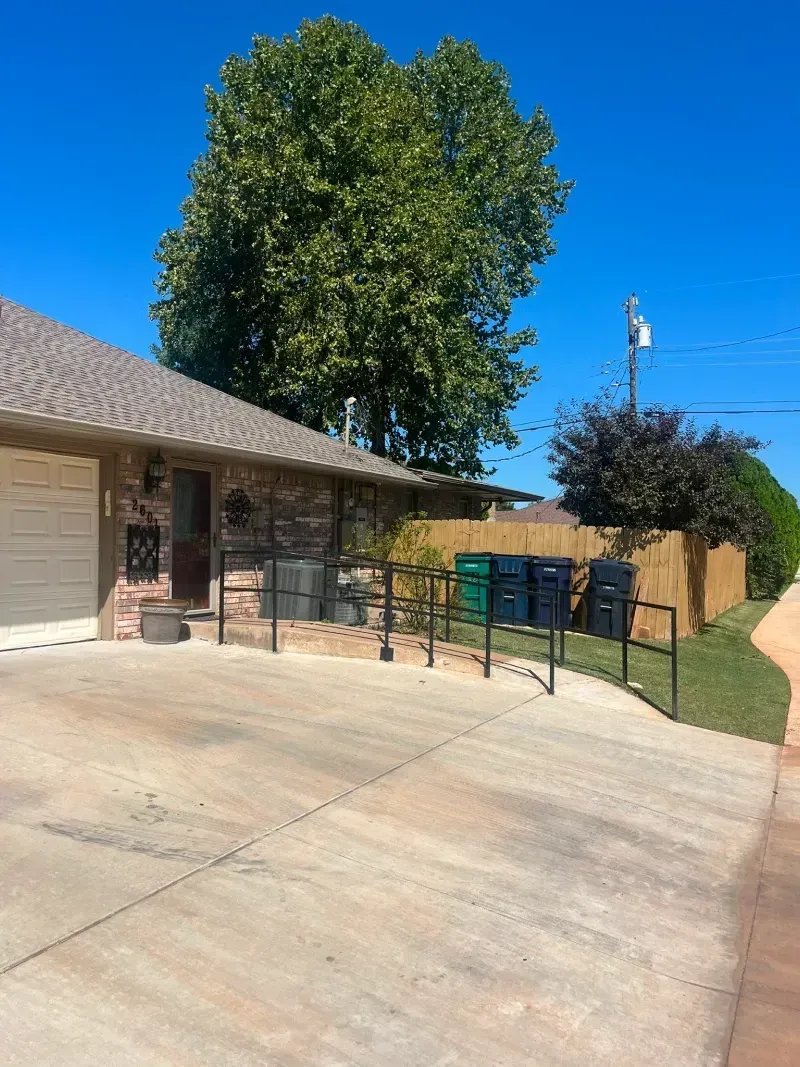 House exterior with concrete driveway, trash cans, wooden fence, and blue sky.
