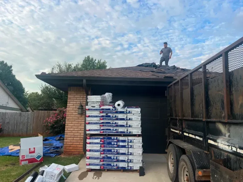 Man on roof tearing shingles with stacked roofing materials in front of garage; trailer nearby.