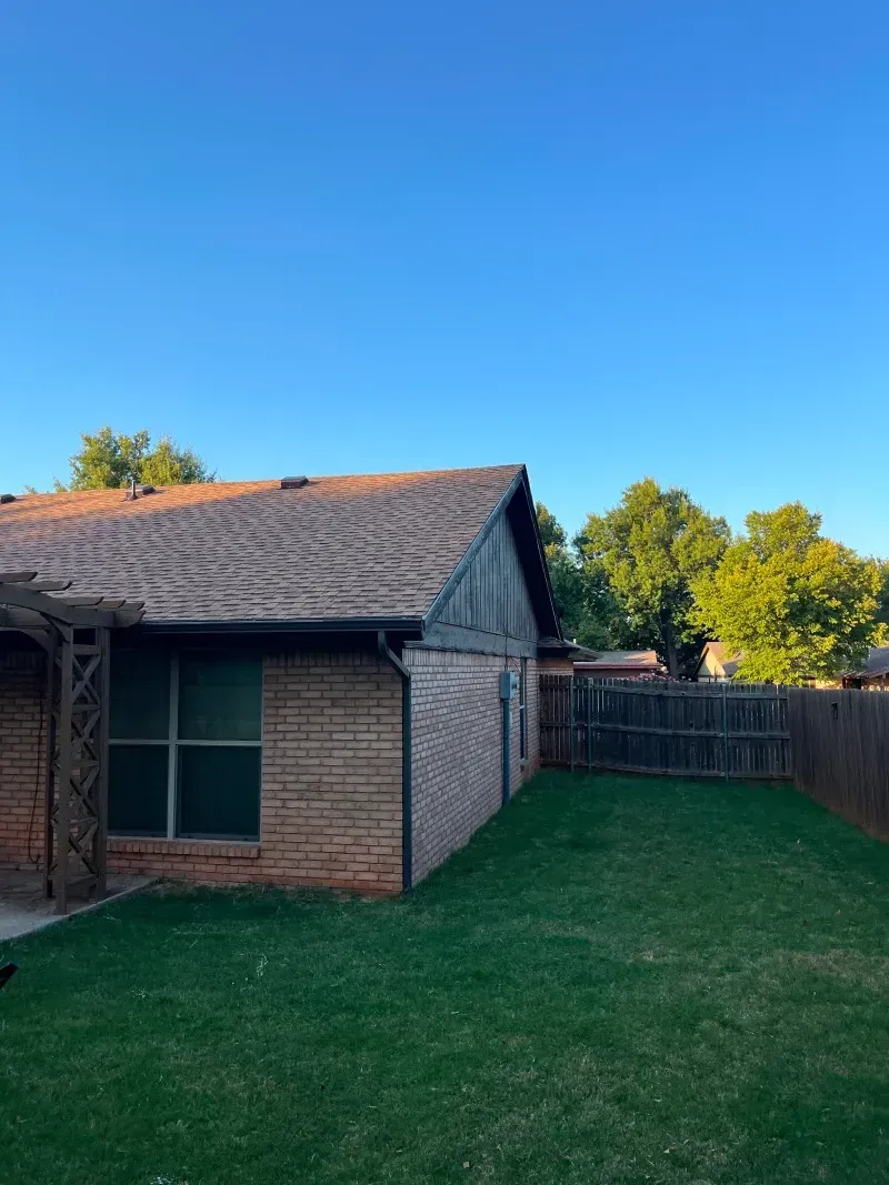 Brick house with a sloped roof, a green lawn, and a wooden fence under a blue sky.