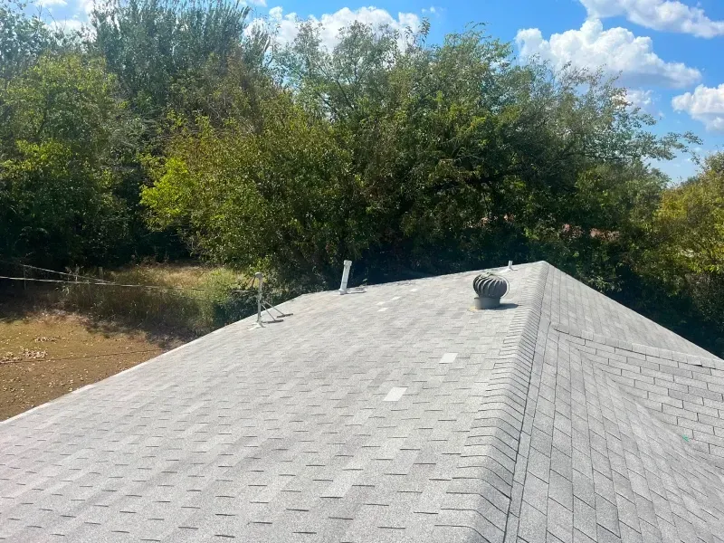 Gray shingle roof of a house with vents, trees in the background, blue sky, and a cloudy day.
