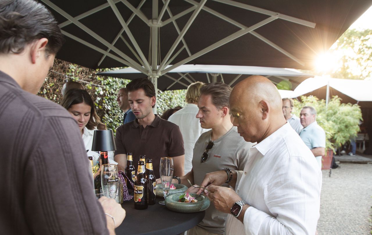 Mensen bij een bijeenkomst in de buitenlucht, rond een tafel met drankjes en eten. Sommigen eten, anderen praten.
