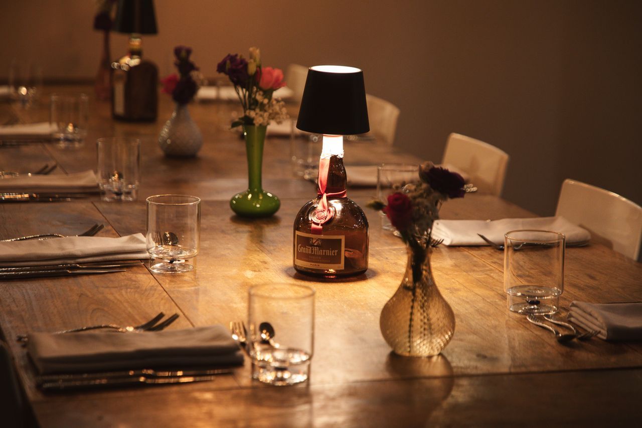 Houten tafel gedekt voor een maaltijd, verlicht door kleine lampjes gemaakt van drankflessen. Vazen met bloemen.