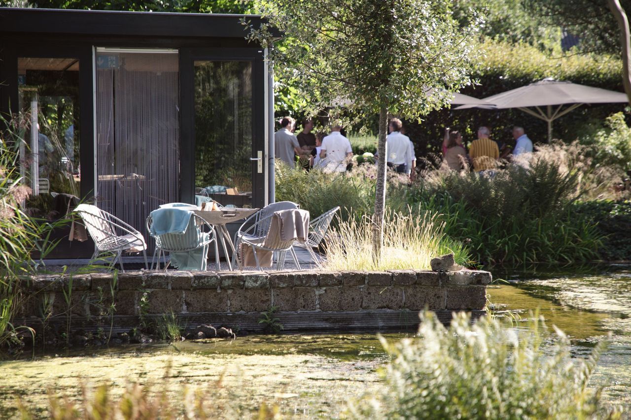 Een tafereel aan het meer met een kleine hut, een tafel met stoelen en mensen die onder een parasol zitten.