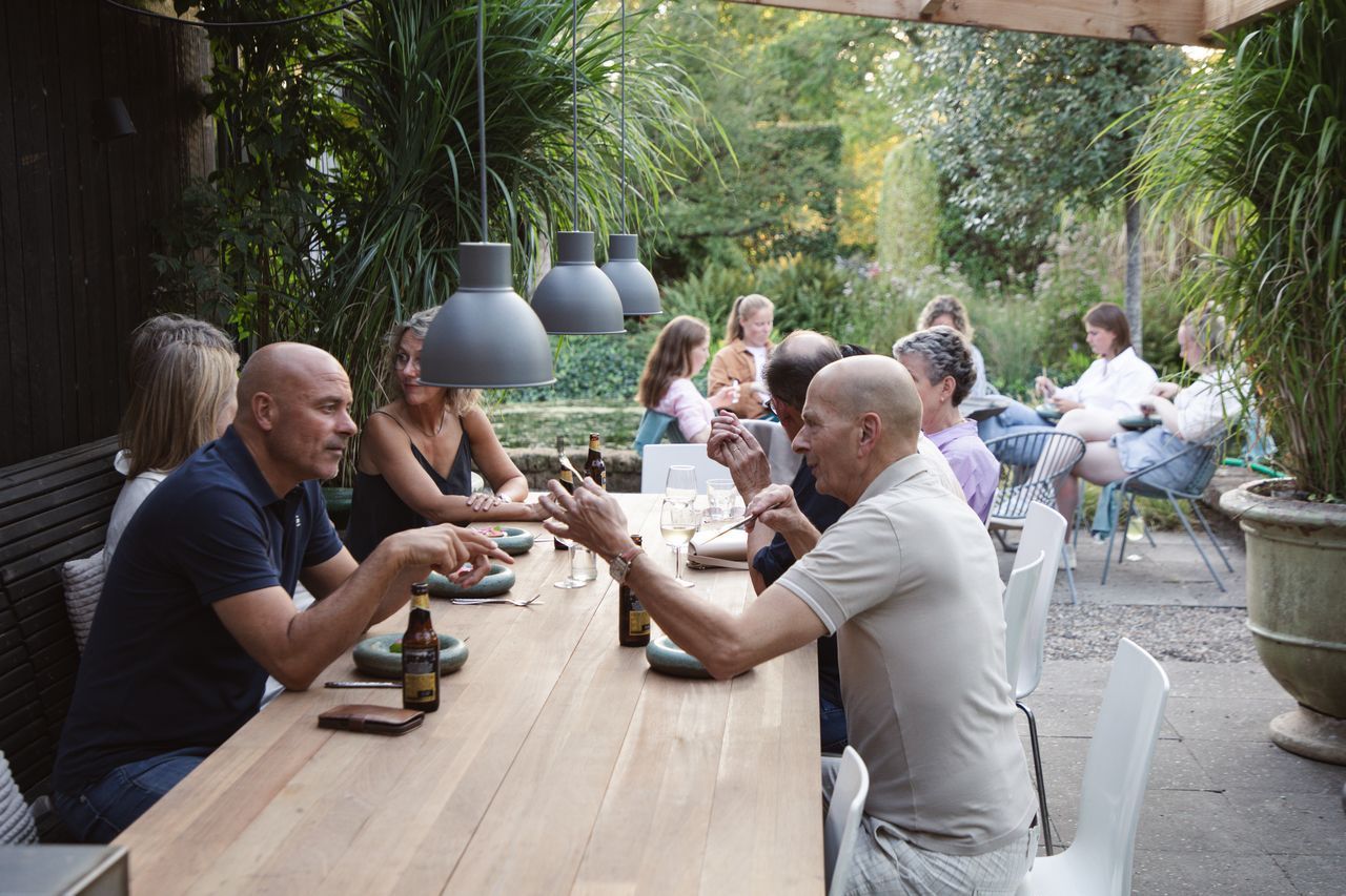 Mensen dineren buiten aan een lange tafel, met groen gebladerte op de achtergrond en grijze hanglampen erboven.