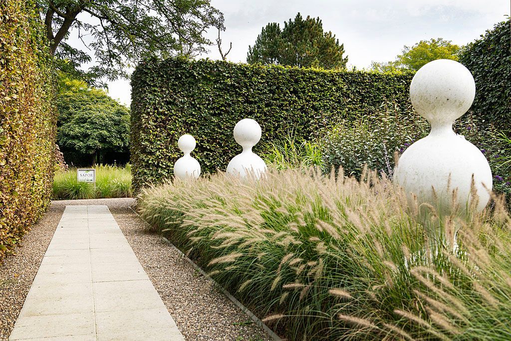 Een tuinpad omzoomd met siergrassen en witte sculpturen, begrensd door hoge groene hagen.