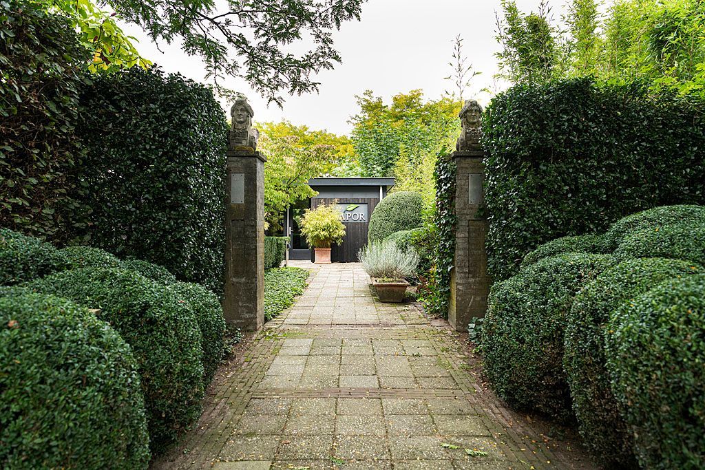 Een stenen pad, omzoomd door hagen en pilaren, leidt naar een donker gebouw.
