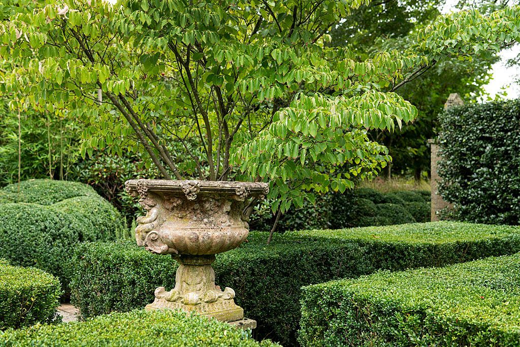 Stenen urn in een formele tuin, omgeven door keurig gesnoeide groene hagen en bomen.
