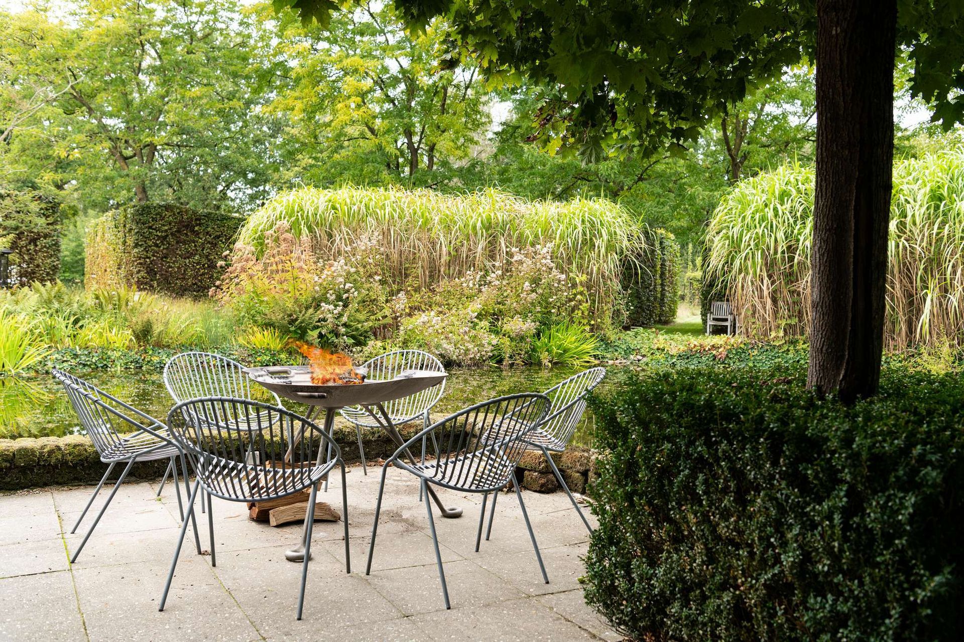 Een zitgedeelte buiten met een barbecue en stoelen bij een vijver, omgeven door weelderig groen en bomen.