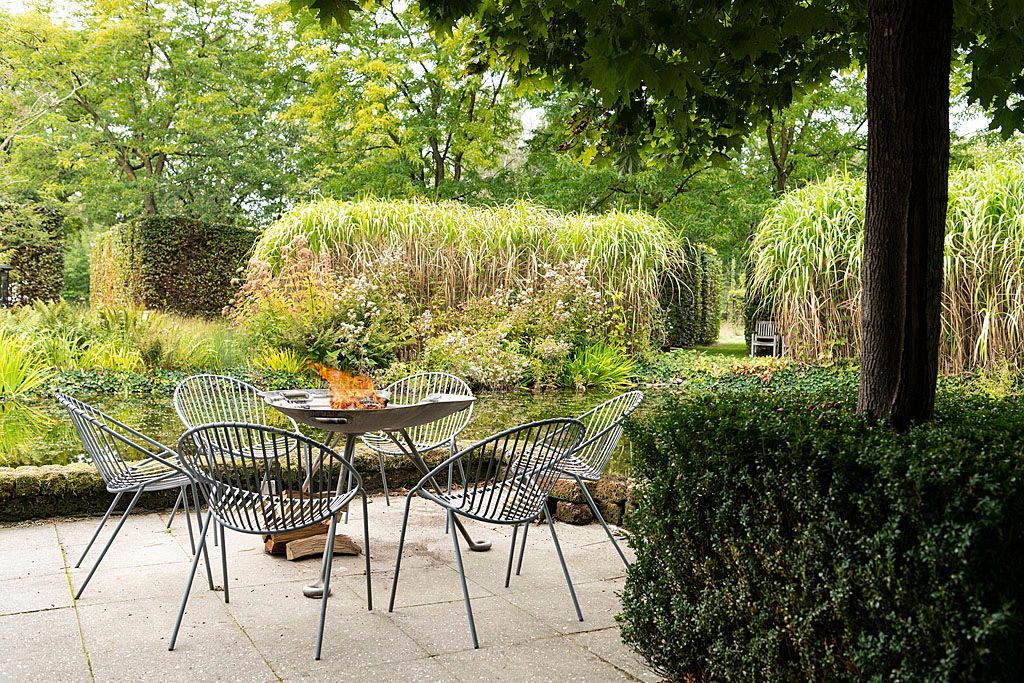 Terrasmeubilair onder een boom in een tuin met keurig gesnoeide hagen en bomen.