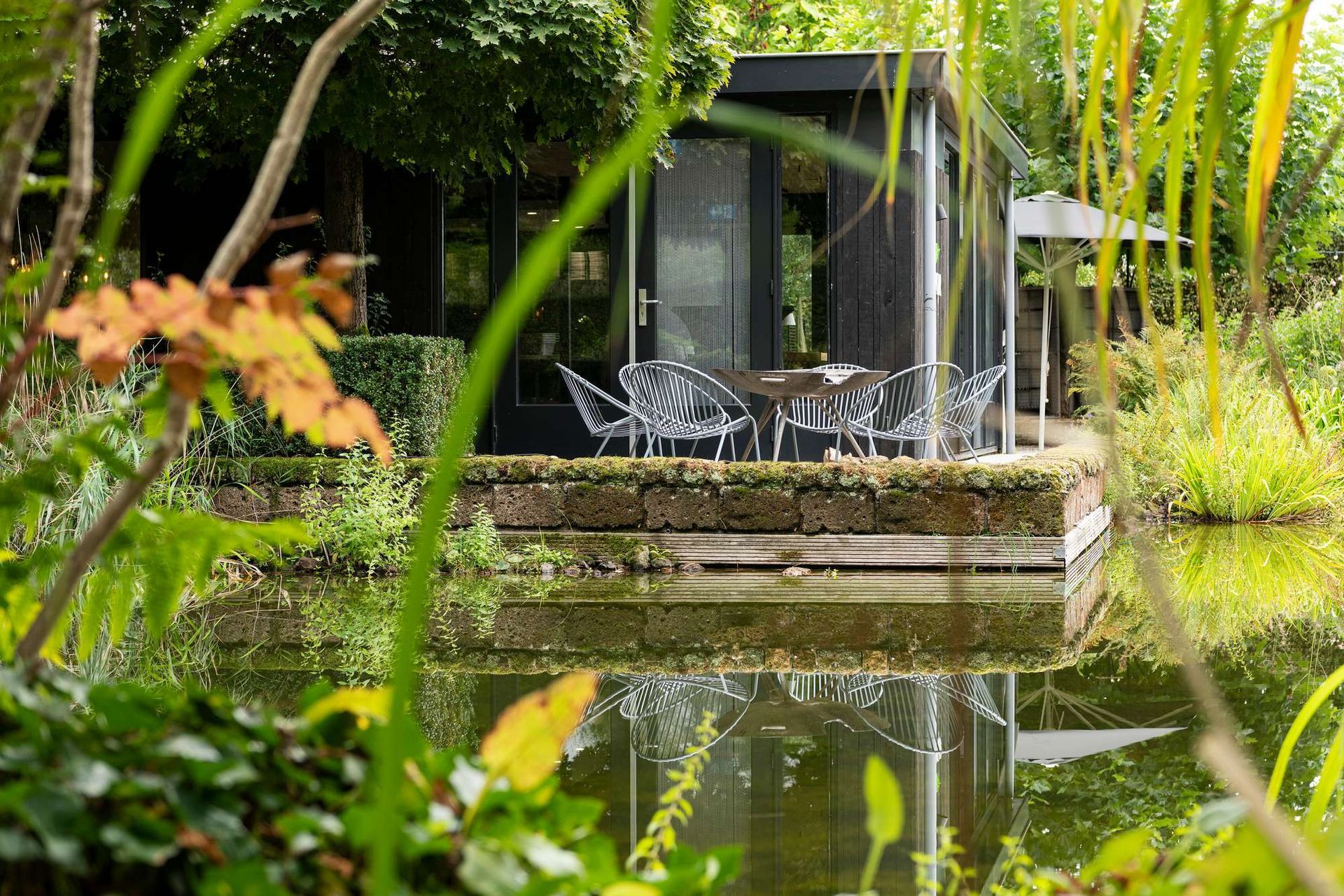 Moderne zwarte hut met glazen wanden en transparante stoelen op een platform boven een vijver.