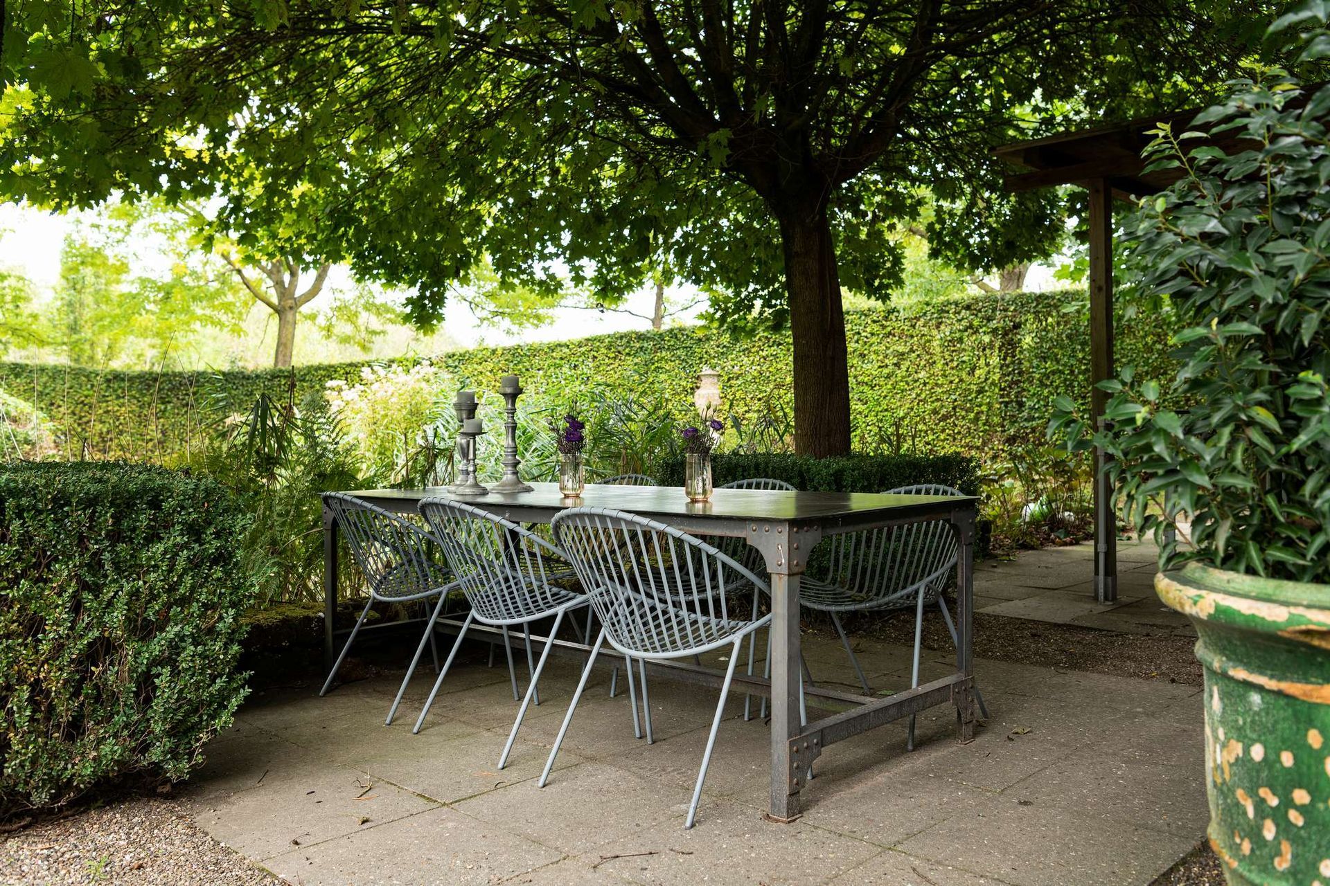Eettafel en stoelen buiten onder een boom, omgeven door groen en hagen.