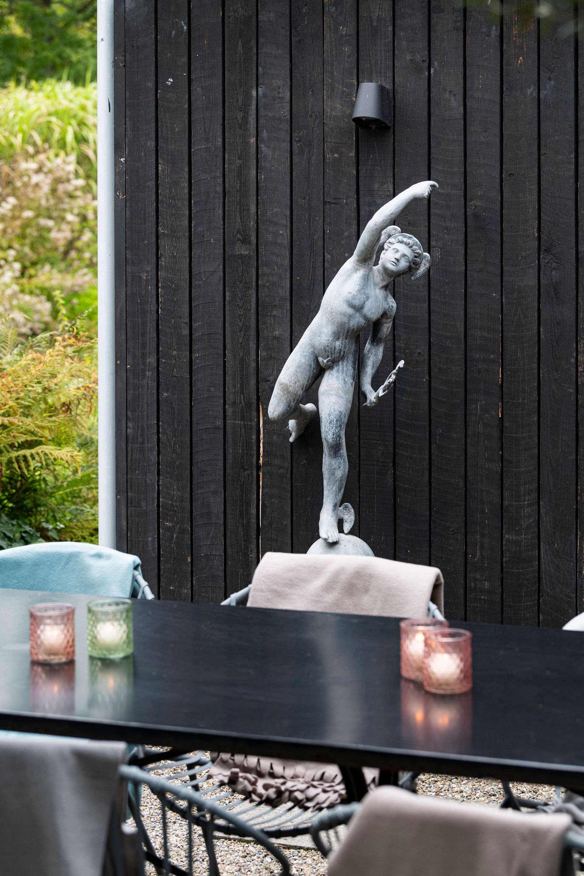 Standbeeld van een vrouw tegen een zwarte houten muur, tafel met kaarsen, buitenomgeving.