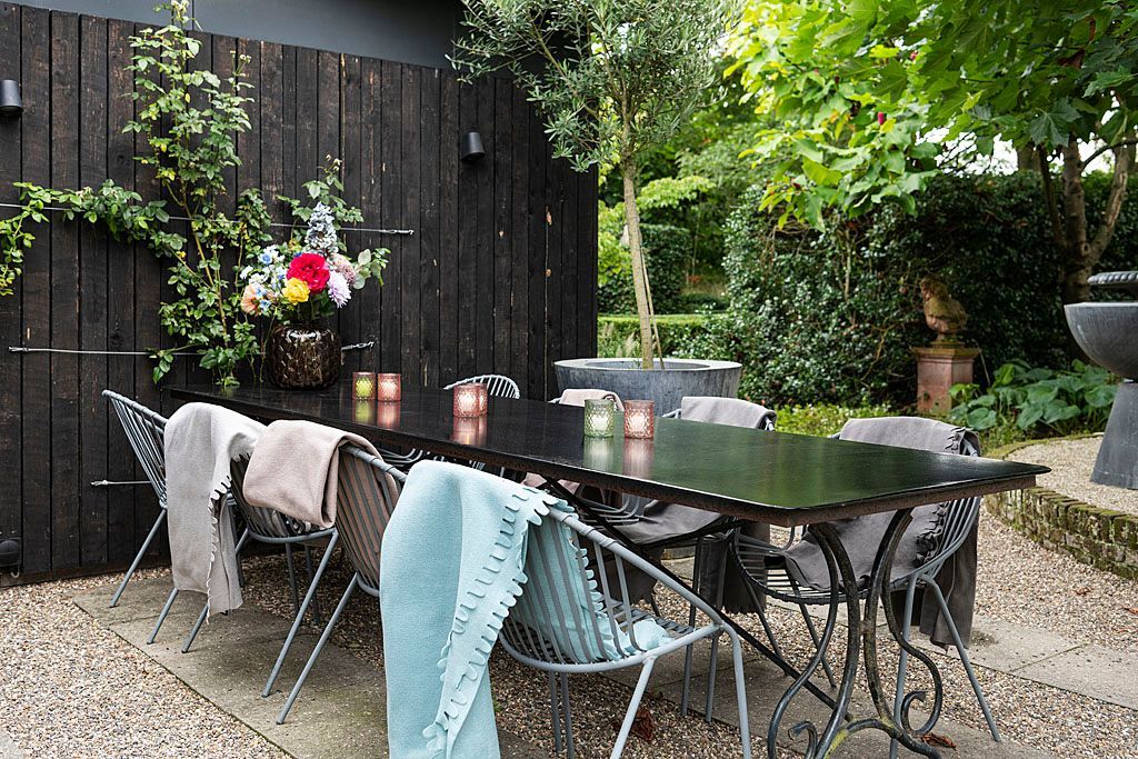 Eettafel voor buiten met stoelen, zwarte muur als achtergrond, planten en decoratieve kaarsen.