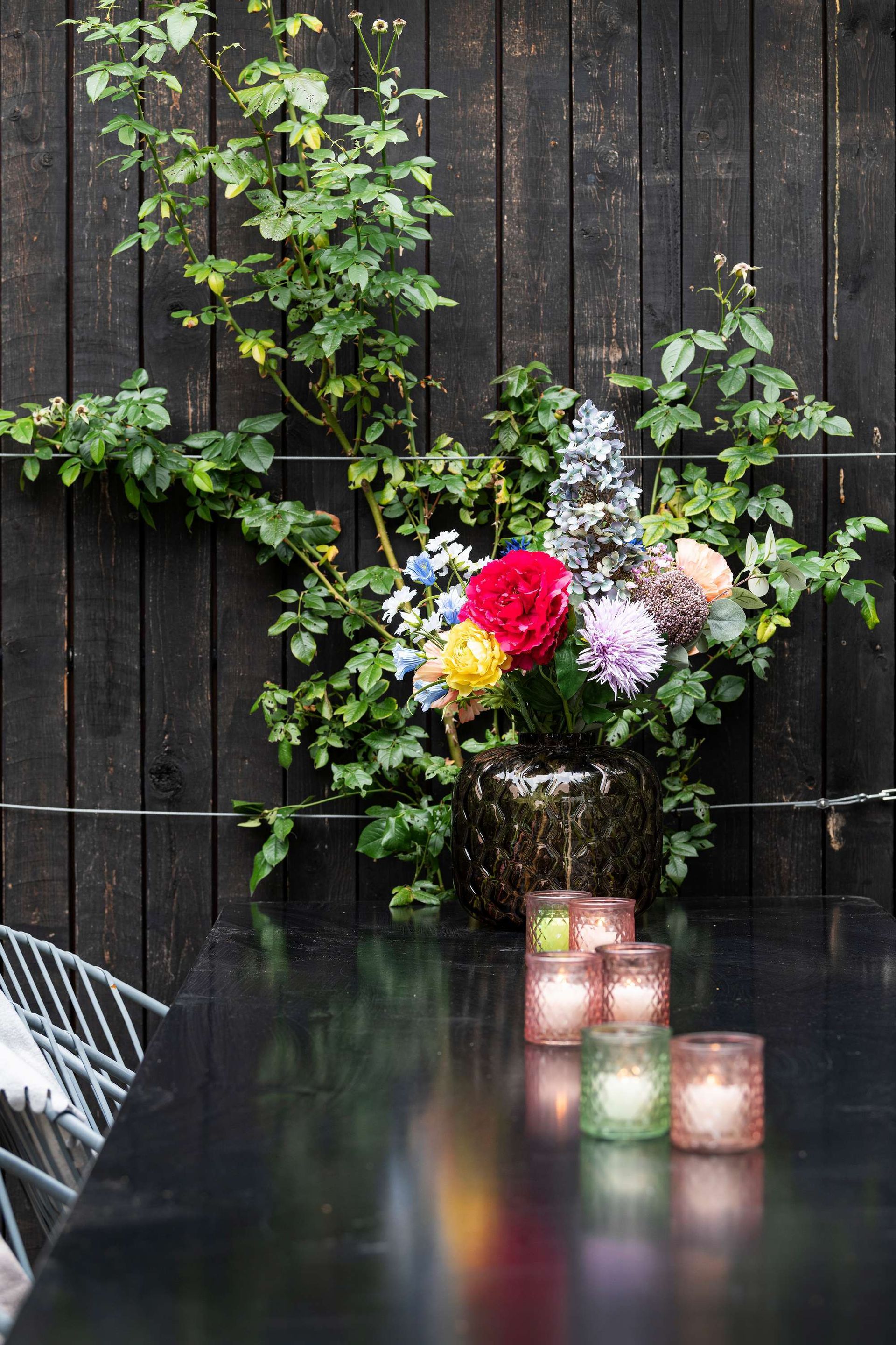 Zwarte tafel met bloemen in een vaas, brandende kaarsen en een klimplant tegen een houten schutting.
