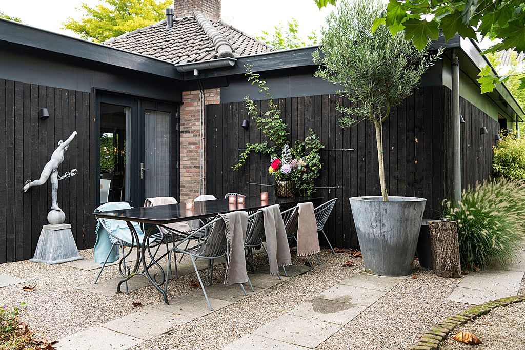 Zwartgekleurd huisje met eethoek buiten, olijfboom in een grote pot en stenen terras.