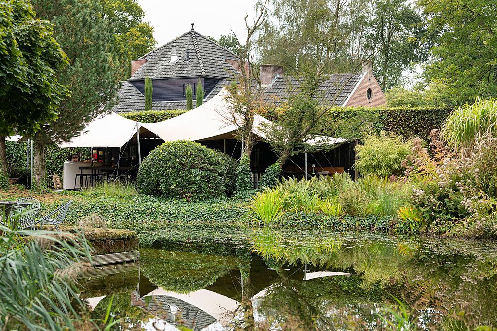 Een vijver met weelderig groen, die een gebouw weerspiegelt met canvas luifels en een donker, schuin dak.