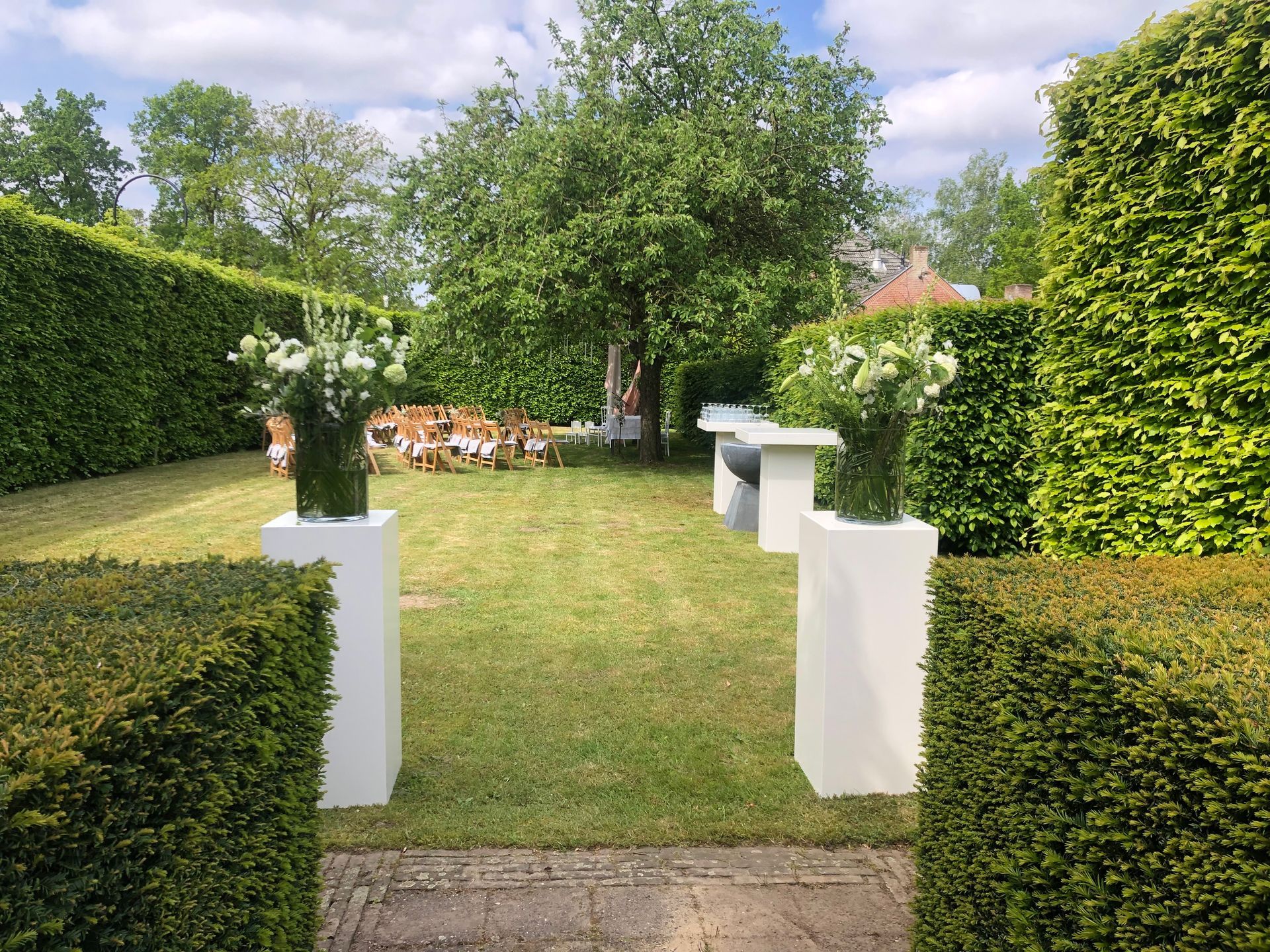 Een weelderige tuin met strak gesnoeide hagen, witte bloemstukken op sokkels en tafels, en een boom onder een blauwe hemel.