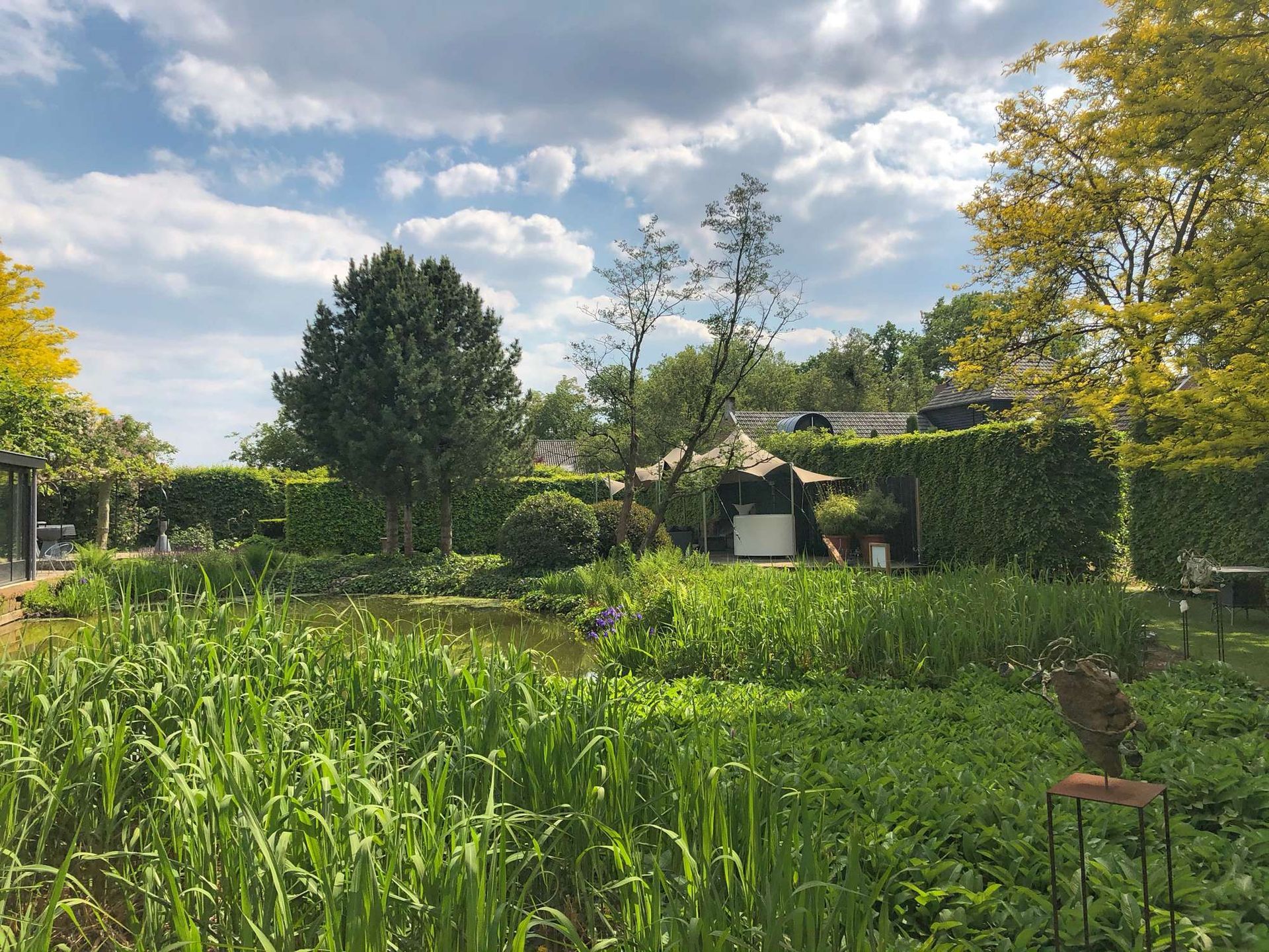 Een weelderige tuin met vijver, hoog gras, hagen en bomen onder een gedeeltelijk bewolkte hemel.