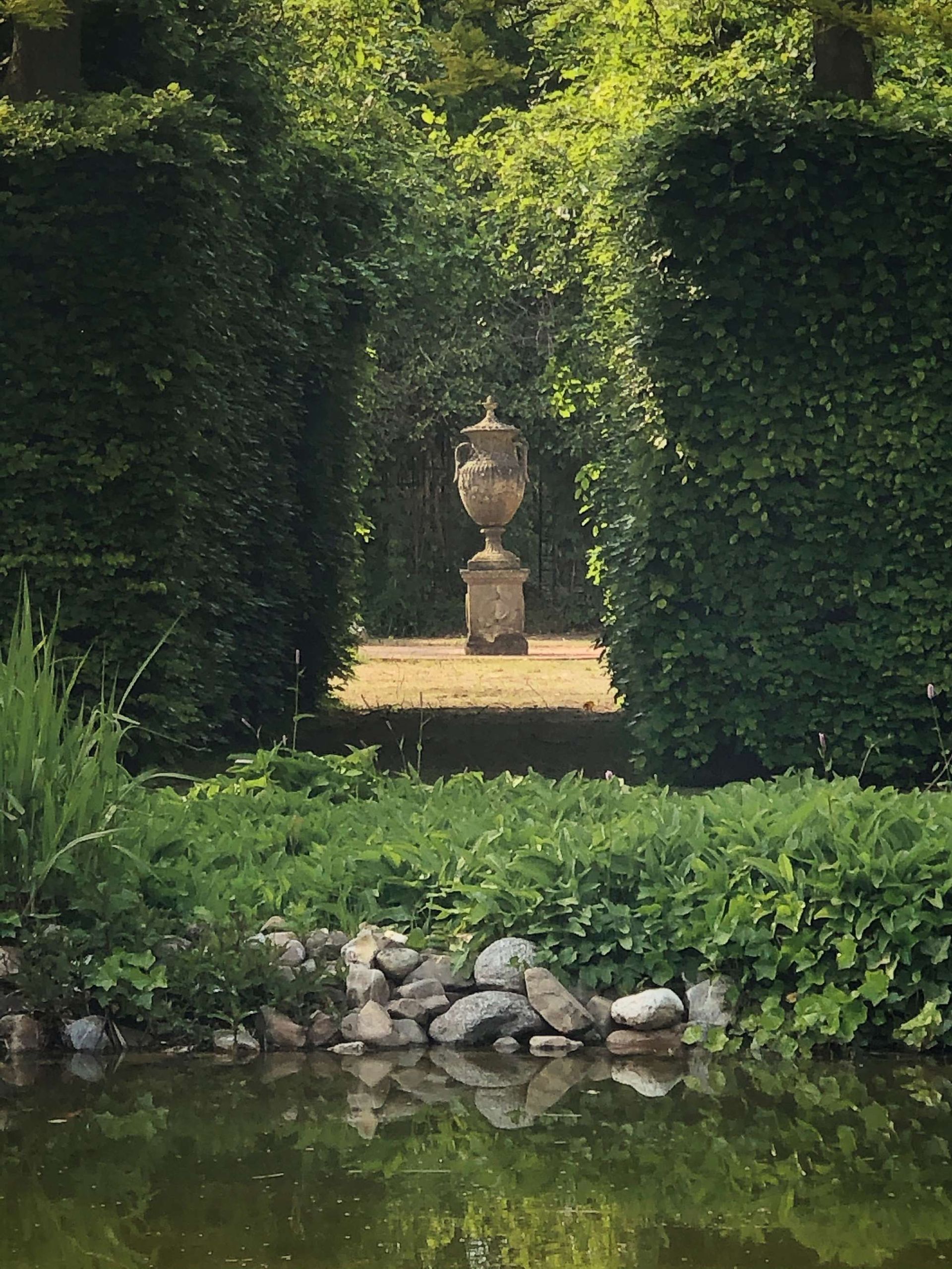 Een formele tuin met een standbeeld dat door een opening in de heg steekt, gezien vanaf een spiegelvijver.